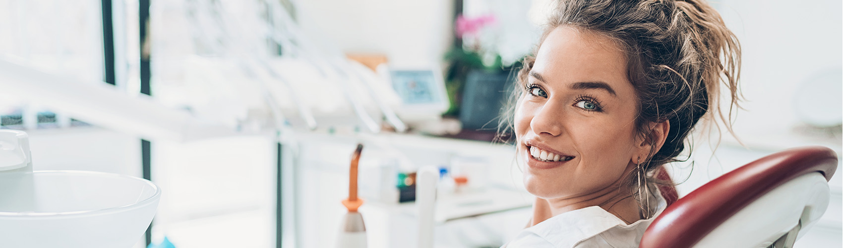 Woman with dark curly hair site in dental chair looking left at the camera smiling